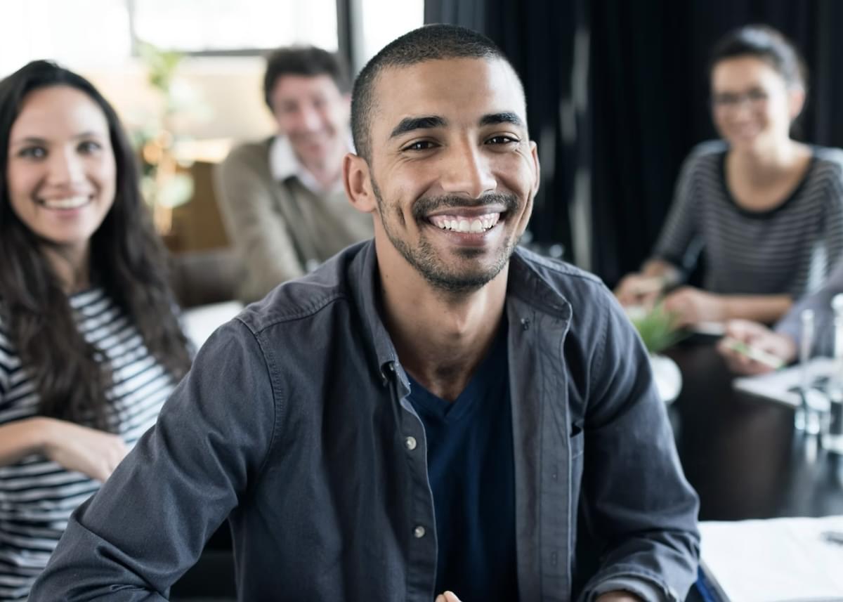 Homem sorridente em primeiro plano, vestindo camisa cinza, acompanhado por colegas de trabalho desfocados ao fundo em um ambiente de escritório.